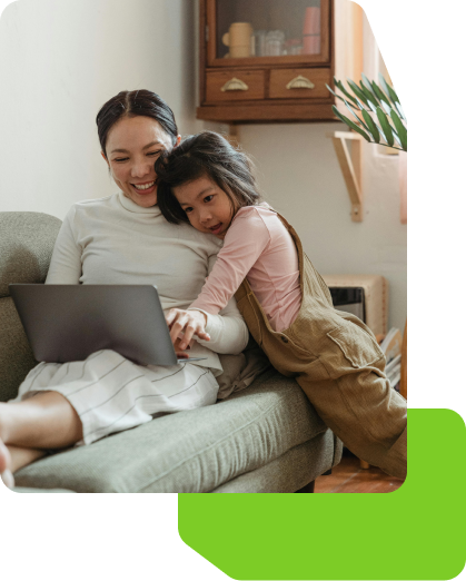 A young child embraces her mother as they smile and look at a laptop together on a couch.