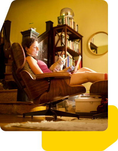 A woman lounges with a laptop and her cat in a cozy, dimly lit room.