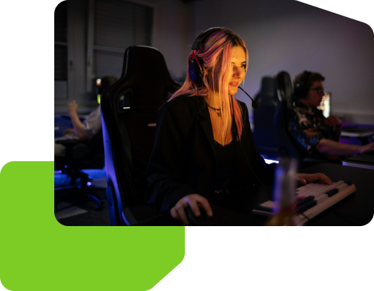 A woman is gaming at a computer setup, wearing a headset, with others playing in the background